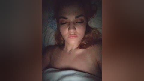 A video shows a fair-skinned woman with reddish-brown hair lying on a bed, eyes closed, wearing a white blanket, and a thin necklace. The background is blurred, emphasizing her peaceful expression.