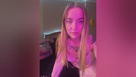 Media: Video of a young, light-skinned woman with long blonde hair, wearing a white top, tattoos on her chest, posing on a bed with pink lighting, blurred background.