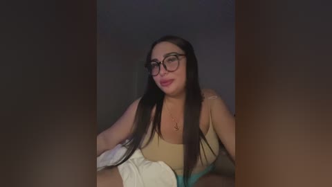 Video of a smiling woman with long dark hair, glasses, and a beige top, sitting on a bed with a white blanket, against a dimly lit background.