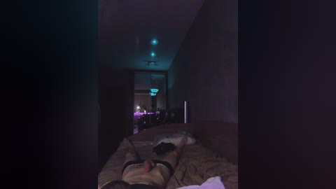 A dimly-lit video of a bedroom at night, showing a person lying on a bed with messy sheets, a partially visible TV, and a chandelier.