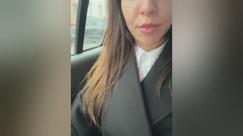 Media: Video of a woman with long, straight brown hair and light skin, wearing a black blazer over a white shirt, sitting in a car. The blurred background shows a cityscape.