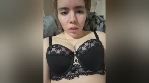 Video of a young woman with fair skin and light brown hair, wearing a black lace bra with floral detailing. She has a neutral expression, sitting on a gray pillow in a dimly lit room.