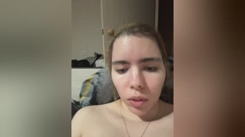 Video of a young, fair-skinned, topless woman with light brown hair in a messy bun, wearing a thin gold necklace. She has a neutral expression. Background shows a messy bedroom with a bed and scattered clothes.