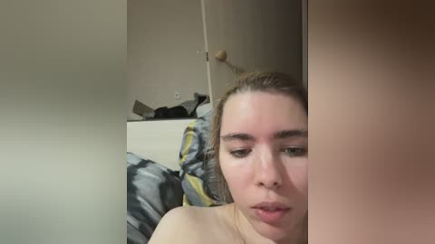 Video of a young woman with light skin and light brown hair, partially obscured by a vertical mirror, lying on a bed with a patterned gray and yellow blanket. Background shows a cluttered room with wooden furniture and scattered objects.