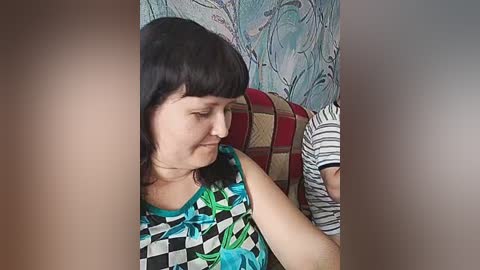 A video of a middle-aged woman with shoulder-length black hair, wearing a colorful, geometric-patterned blouse, sitting on a patterned, red and brown cushion. The background features a floral-patterned wall in pastel colors.