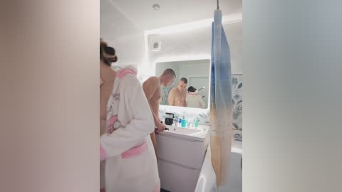 Media: A video of a modern bathroom with a white-tiled wall, a white bathtub, and a man in a white robe with pink trim, drying his hair.