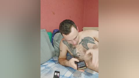 Video of a shirtless man with a large, intricate tattoo on his chest, lying on a bed with a blue and white checkered blanket. He is holding a smartphone, and there's a cellphone on the bed beside him. The room has red and peach walls.