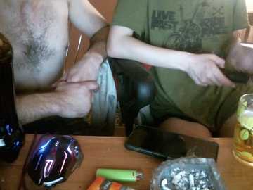 Media: Video of two shirtless men, one with a beer belly, smoking a cigarette while the other holds a lighter. Background shows a cluttered wooden table with a glass, lighter, and cigarettes.