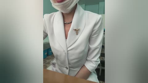 Media: A video of a woman in a white nurse's uniform, wearing a face mask and a butterfly brooch, standing in a medical setting with cabinets in the background.