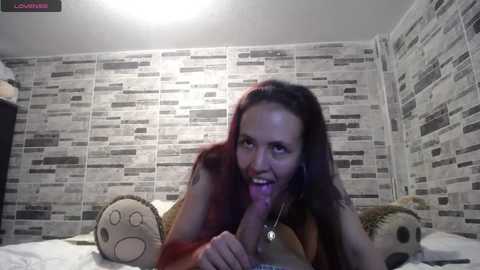 Video of a young woman with long dark hair, fair skin, and small breasts, wearing a brown top, lying on a bed with a gray and white striped wallpaper background. She is licking a large, erect penis.