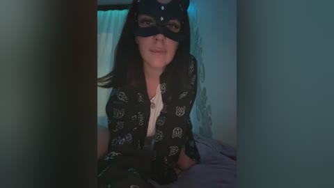 Video of a woman with long dark hair and fair skin wearing a black mask, black shirt with white pentagram pattern, and a necklace, seated on a bed in a dimly lit room.