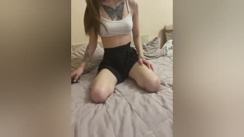 Media: Video of a young woman with fair skin and long brown hair, kneeling on a beige bedspread. She wears a white crop top revealing a butterfly tattoo on her chest and black shorts. The background features a beige wall and a partially visible pillow.
