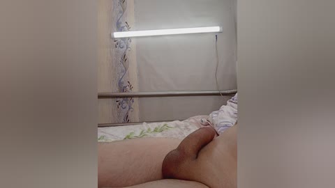 Media: Video of a man lying on a bed, nude, with a semi-erect penis visible. He is in a hospital room with a beige curtain and a fluorescent light overhead.