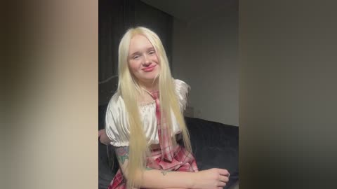 Media: Video of a smiling young woman with long, straight blonde hair, wearing a red and white plaid dress, sitting on a dark couch in a dimly lit room.