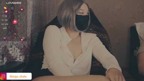 A video of a light-skinned woman with a bob haircut, wearing a black face mask and a white blouse, sitting in a dimly lit room.