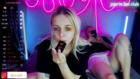 Video of a blonde woman with bright eye makeup and tattoos, holding a black phone, in a dimly-lit room with neon lights.