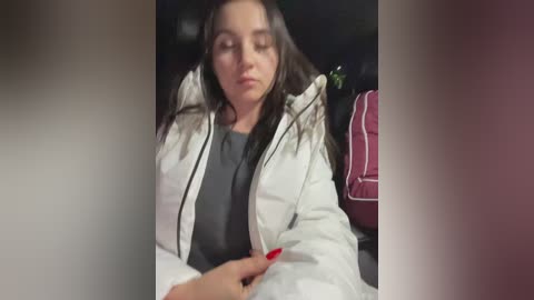 Media: A video of a young woman with long brown hair, closed eyes, and a neutral expression, wearing a white jacket and a gray shirt, sitting in a car with a red seatbelt visible.