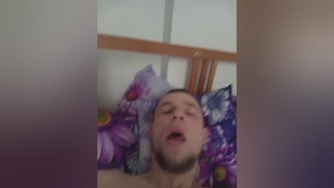 Video of a bearded, fair-skinned man lying on a bed with floral-patterned pillows, mouth open in a relaxed expression, head slightly tilted back.