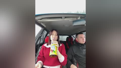 Video of a woman in a red sweater drinking a soda through a straw, sitting next to a man in a black jacket in a car with a sunroof, blurry background.