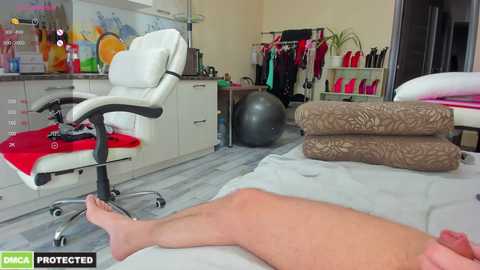 Media: Video of a modern, well-lit spa room with a white leather massage chair, a gray massage table, and a person's legs visible on the table. The background features shelves with various spa products and a colorful fish tank.