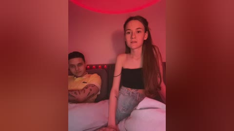 Media: Video of a young, slim, light-skinned woman with long brown hair, wearing a black crop top and high-waisted jeans, sitting on a bed under red lighting, next to a man in a yellow shirt.