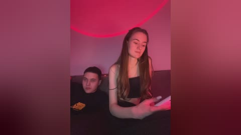 Media: Video of a young man in a black hoodie with a yellow logo, sitting on a couch, and a young woman with long brown hair, wearing a black crop top, playing a video game. The room is dimly lit with red and pink lighting.