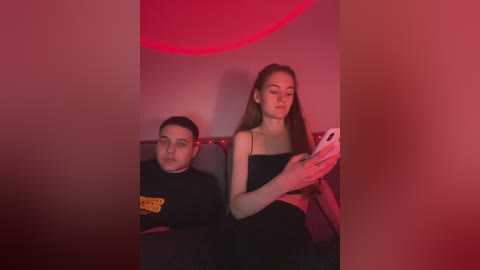 Media: Video of two young people under red light: a boy in a black T-shirt, seated, and a girl in a black crop top, standing, holding a phone.