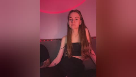 Media: Video of a young woman with long brown hair, wearing a black crop top and high-waisted pants, seated on a grey couch in a dimly lit room with red lighting.