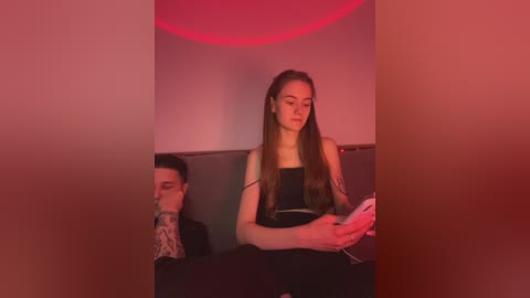 Media: Video of a young woman with long brown hair, wearing a black top, seated next to a man with a tattooed arm. They're in a dimly lit room with a red light, holding a phone.