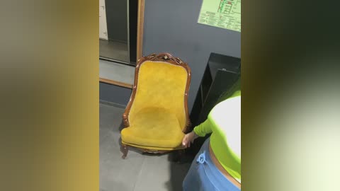A video of a vintage yellow velvet chair with carved wooden arms, placed in a modern room with a green wall and a black table. A person in a green shirt and blue pants stands beside the chair.