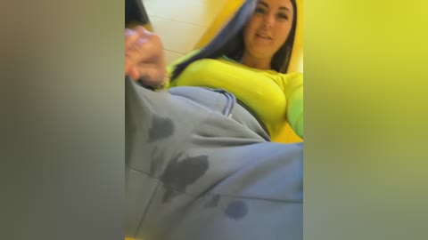 Video of a young woman with medium-length dark hair, wearing a yellow shirt and gray sweatpants, seated in a yellow chair.