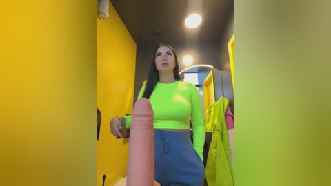 Video of a woman with long black hair, wearing a neon yellow crop top and blue skirt, standing in a yellow hallway, holding a large, erect penis.