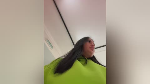 A video of a woman with long, straight black hair, wearing a bright green cape, standing in a white-walled room with a ceiling light.