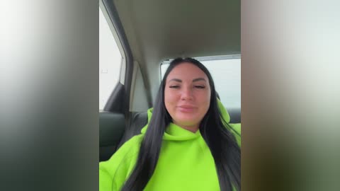 A video shows a young woman with long black hair, fair skin, and closed eyes, wearing a bright neon green hoodie, sitting in a car. The background is blurry, and the car's interior is dimly lit.