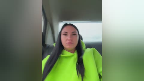 A video of a woman with long black hair wearing a bright neon green hoodie, seated in a car, with blurry vehicle interior and blurred background.