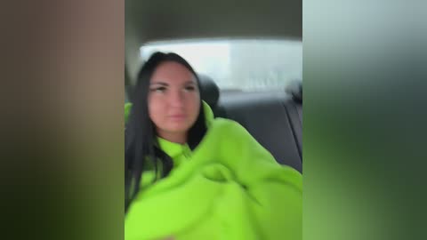 Video of a woman with long black hair wearing a bright neon green hoodie, driving a car with blurred background.
