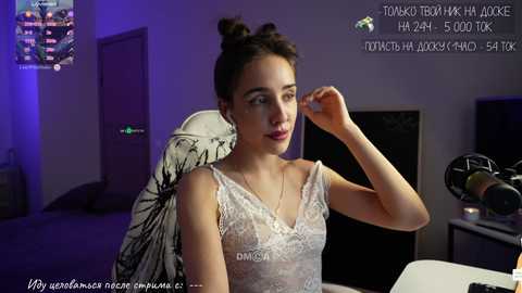 Media: A video of a fair-skinned woman with dark hair in a bun, wearing a sheer lace top, sitting in a gaming chair with headphones, in a dimly lit room with gaming equipment.