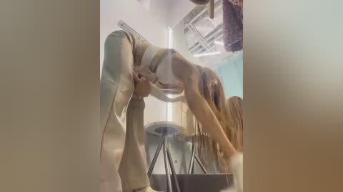 Media: A video captures a woman in a beige crop top and high-waisted pants bending over a metal table in a dimly lit room with a teal wall and industrial ceiling.