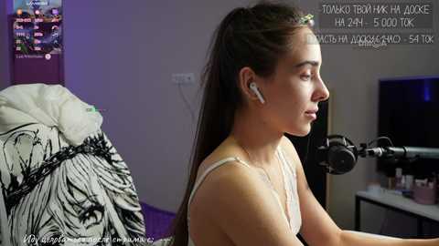 Media: A video of a young woman with light skin and long brown hair, wearing a white tank top, sitting at a desk with a black and white floral blanket, headphones on, recording herself.