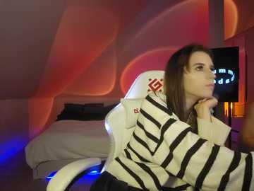 Media: Video of a young woman with straight brown hair, wearing a striped sweater, sitting in a white gaming chair in a dimly lit room with red and blue ambient lighting.