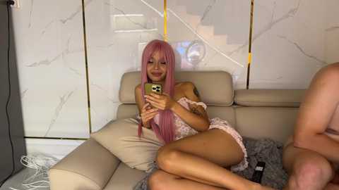 Media: Video of a young, slender woman with long, pink hair, wearing a floral dress, sitting on a beige couch, smiling while taking a selfie.