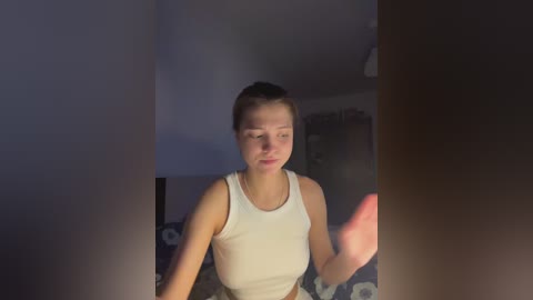 A video of a young woman with short brown hair, wearing a white tank top, standing in a dimly lit room with a dark bed and a blurred background.