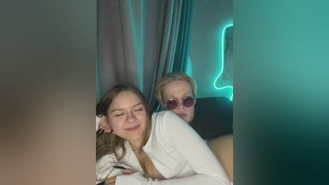 Video of two young women, one with long brown hair and the other with blonde hair, wearing white tops, lounging together in a dimly lit room with teal neon lights and gray curtains.