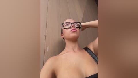 Media: Video of a young woman with light skin, wearing black-framed glasses and a black strapless top, standing in front of a wooden door. She has a confident expression, with one hand on her head.