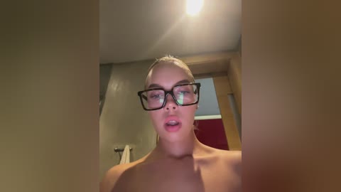 Media: Video of a topless woman with fair skin, wearing black-rimmed glasses, in a bathroom with beige walls, a red door, and a towel rack.