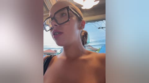 Media: Video of a young woman with light skin, glasses, and brown hair in a bun, wearing a black top, taken indoors near a window with an outdoor view of houses.