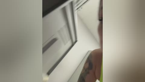 Media: Video of a partially visible woman with shoulder-length blonde hair, wearing a green top, standing in a narrow, dimly lit hallway with white walls and a metallic door.