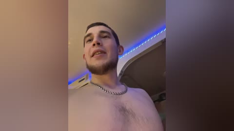 Media: A video of a shirtless man with a trimmed beard and short dark hair, wearing a silver chain necklace, standing in a dimly lit room with blue LED strip lights.