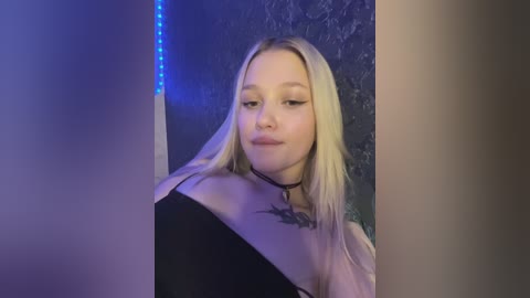 Media: Video of a young, fair-skinned woman with long, straight blonde hair, wearing a black choker necklace and a black top. She stands against a textured, dark blue wall with blue LED lights on the left.