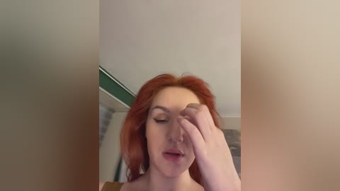 Media: A video of a fair-skinned, red-haired woman with closed eyes, touching her face, in a dimly lit room with beige walls and a green trim.
